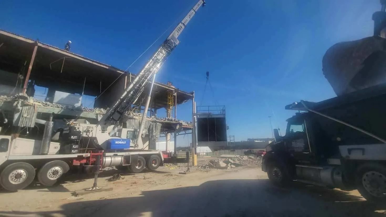 Construction site: Crane demolishing a building; truck loads debris against a bright blue sky.