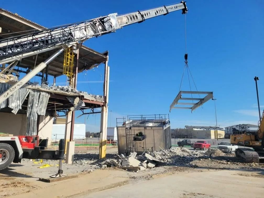 Crane removing metal frame from partially demolished building; rubble on ground; clear blue sky.