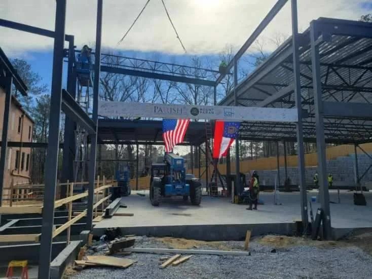 Construction site with steel frame. American and North Carolina flags hang. Workers and a lift are visible.