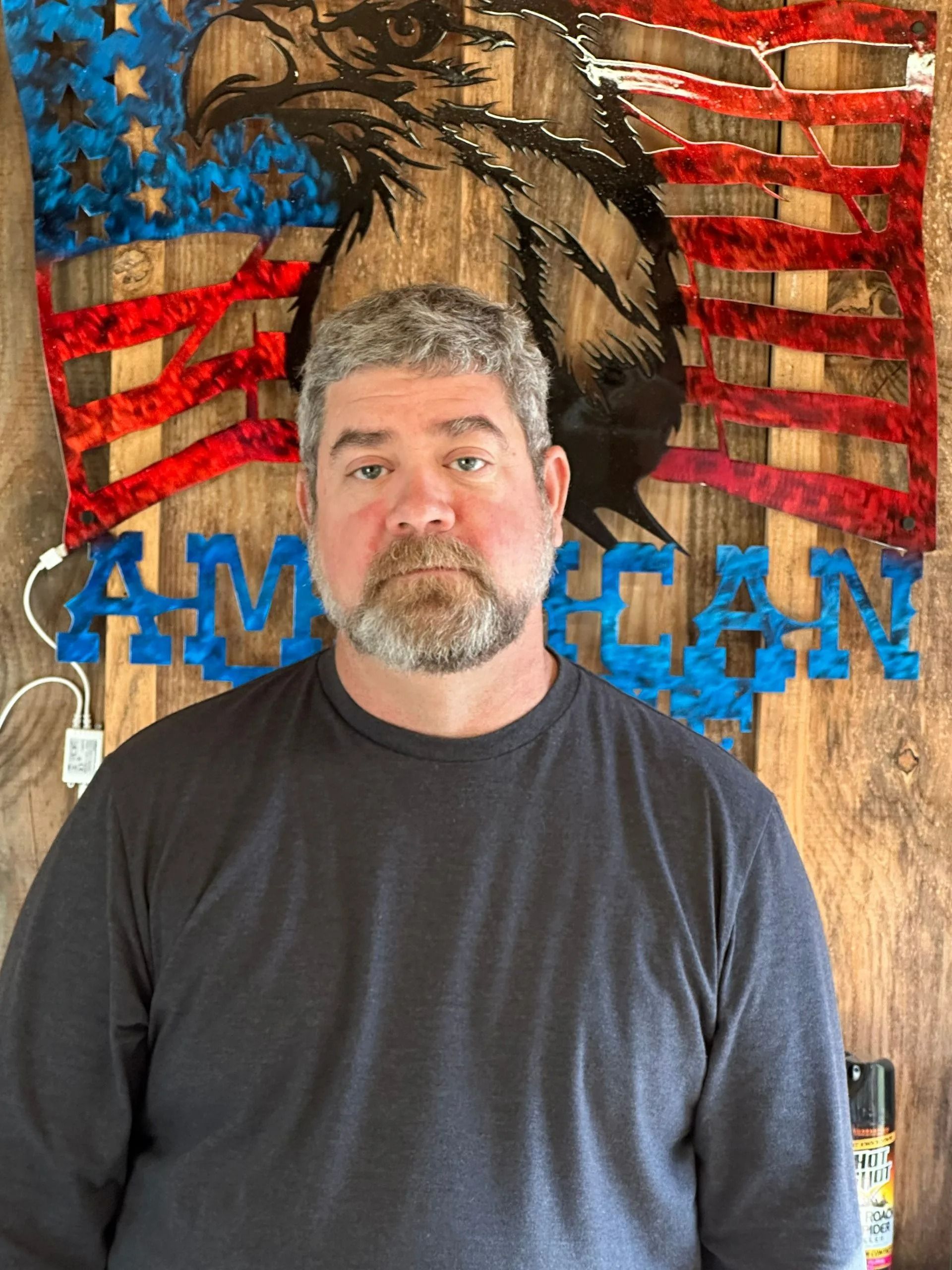 Man with graying beard stands in front of a wood American flag with an eagle carving.