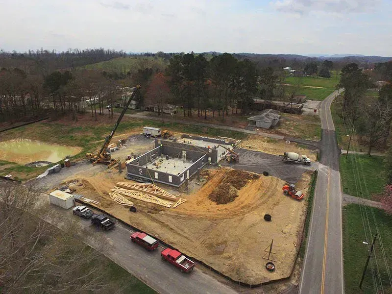 Construction site aerial view; concrete foundation, lumber, heavy machinery, road, and surrounding landscape.