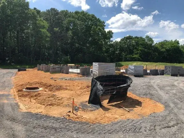 Construction site with gravel, dirt, concrete blocks, and a black mesh drainage structure. Trees in the background, blue sky.