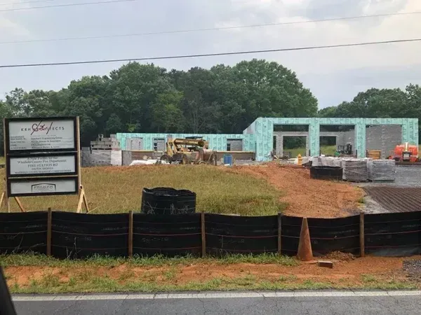 Construction site with framing, surrounded by a black fence and signage, in front of a treeline.