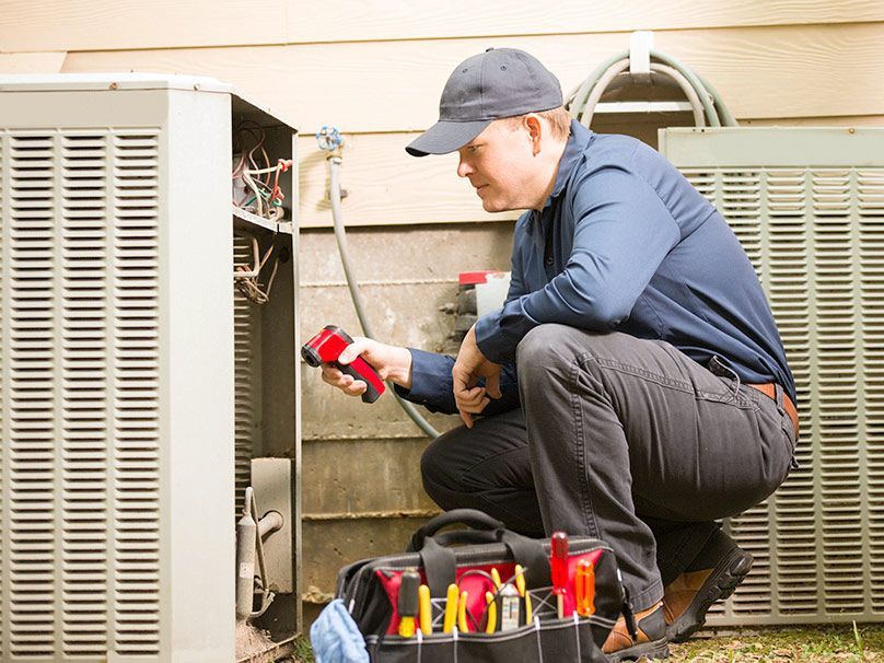 HVAC technician inspecting an outdoor unit. He uses a flashlight, kneeling near his toolbox.