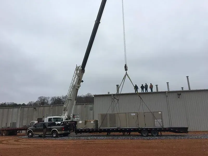 Crane lifting equipment onto a building roof from a flatbed truck; workers observe. Cloudy sky.