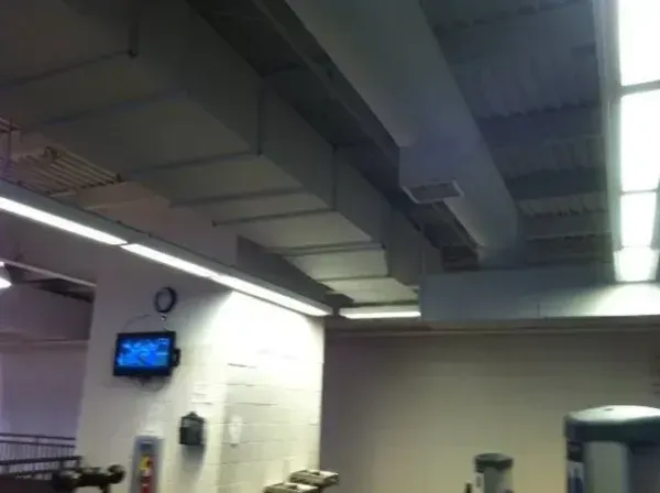 Gym ceiling with rectangular ducts, fluorescent lights, a clock, and a wall-mounted TV.