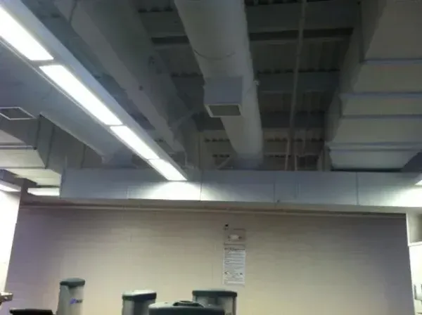 Indoor view of a ceiling with rectangular and circular ventilation ducts, lights, and a white wall below.
