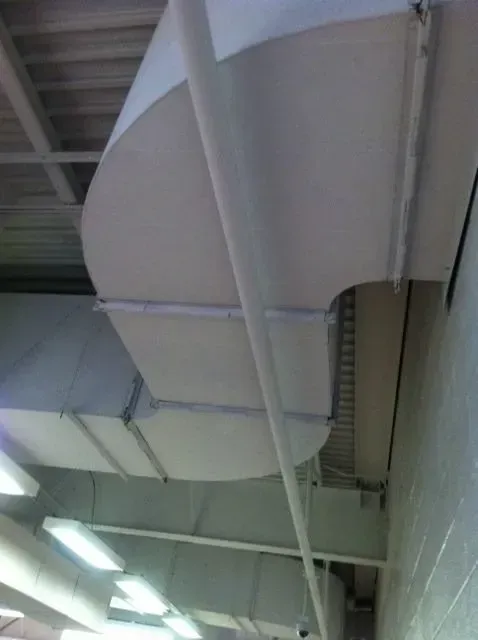 White HVAC ductwork, curved at a ceiling, with support beams and fluorescent lights below.