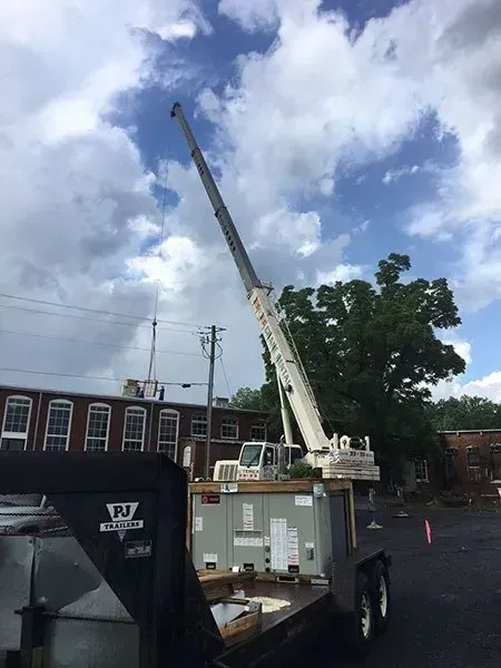 Crane lifting an HVAC unit from a trailer near a building under a cloudy sky.
