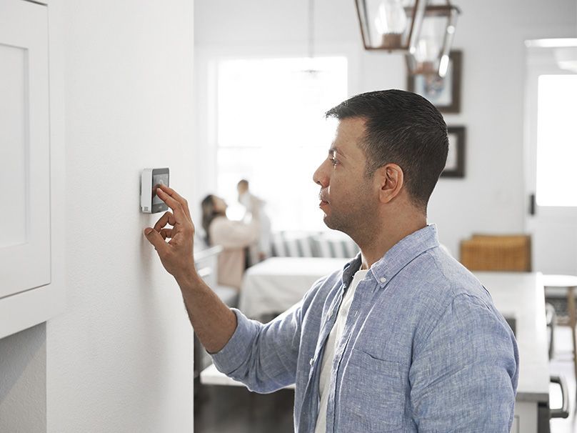 Man adjusts thermostat on a white wall. Soft blue shirt. Family in background.