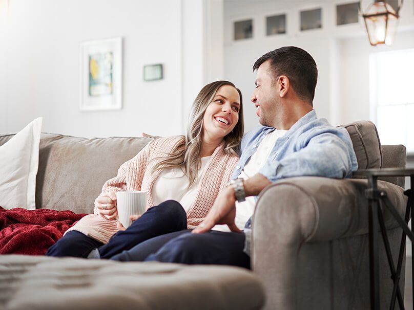Couple smiles at each other on a gray sofa in a living room, woman holding a mug, blanket present.