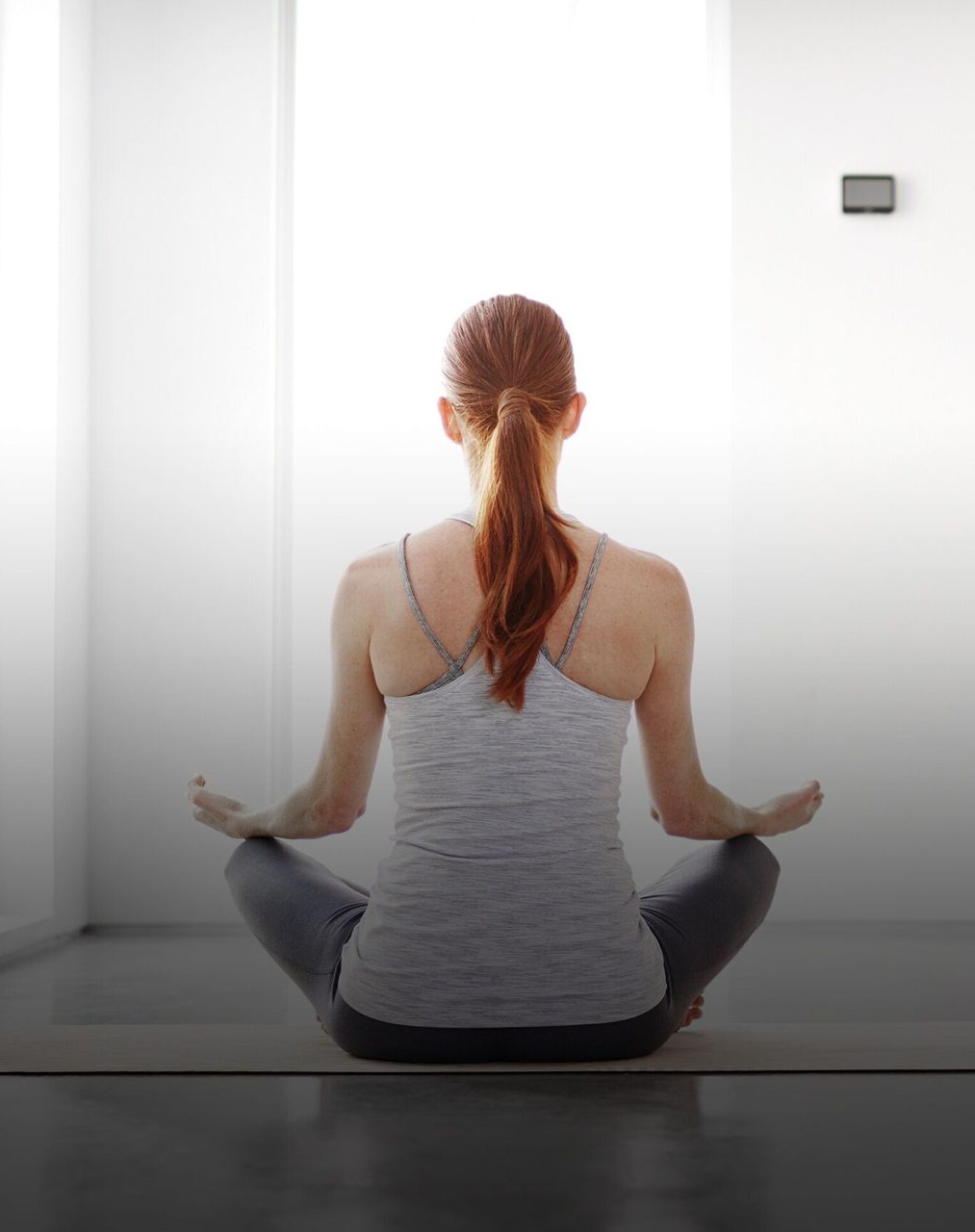 Woman with red hair, in yoga pose on mat. Indoors, facing away, wearing gray top and leggings.