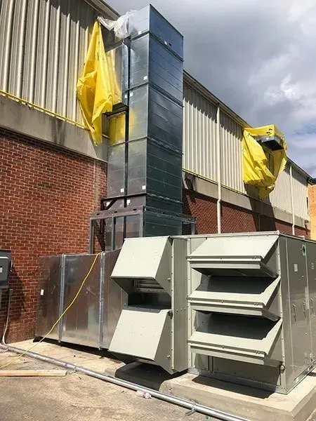 Exterior view of HVAC equipment; silver metal, vents, and a vertical duct system against a brick and white building.