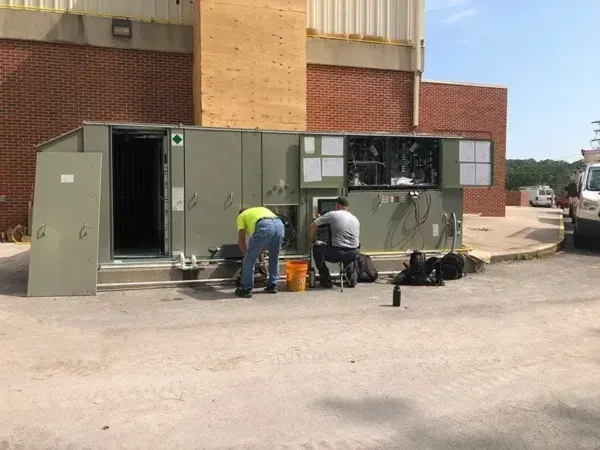 Two men working on a large outdoor HVAC unit outside a brick building.