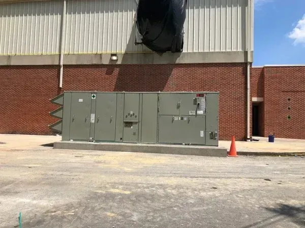 HVAC unit on a concrete base against a brick and metal building. A black tarp is above.