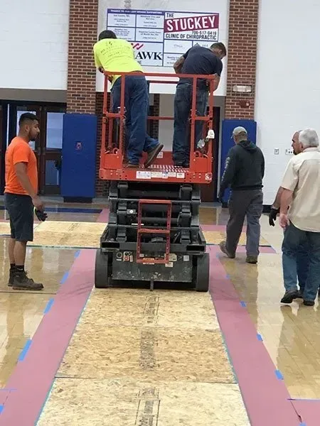 Workers on a lift installing a pink border on a gymnasium floor. Others observe nearby.