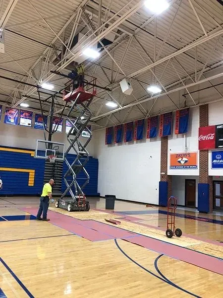 Two workers in a gymnasium. One on a scissor lift near the ceiling, the other on the floor. Basketball court with banners.