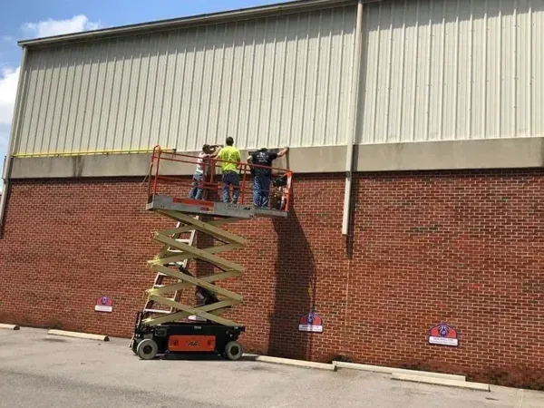 Three people on a scissor lift working on the exterior of a brick building with white siding.