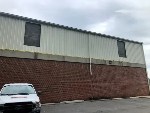 Brick building with a white metal upper section. Two covered windows are visible. White vehicle in the foreground.