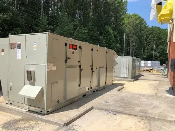 Large, beige HVAC unit outside, next to a smaller, light gray unit. Background trees and a building.