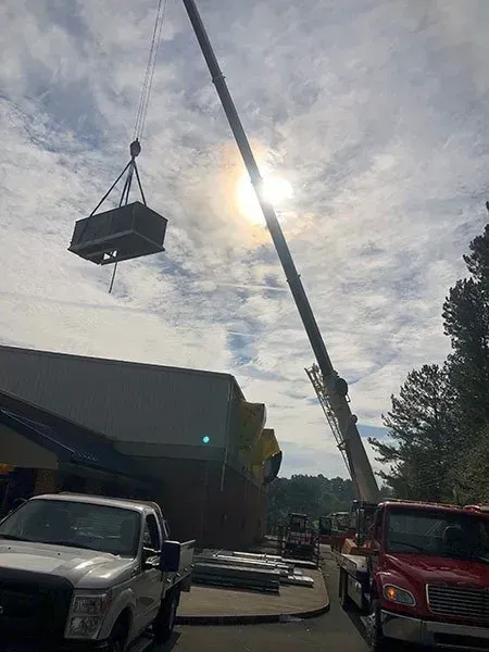 Crane lifting an HVAC unit onto a building under a cloudy sky. Trucks and equipment are on the ground.