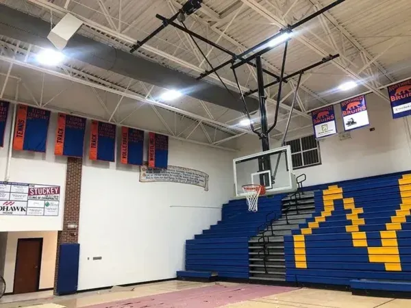 Interior view of a school gymnasium with blue and gold bleachers, a basketball hoop, and banners.