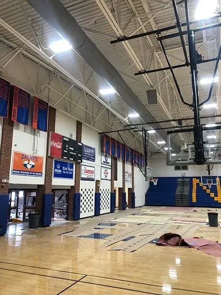 Gymnasium interior with unfinished wooden floor; banners and scoreboard on brick wall.
