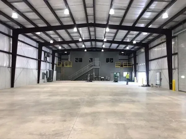 Interior of a large, empty metal warehouse with concrete floor, overhead lighting, and a small office area.