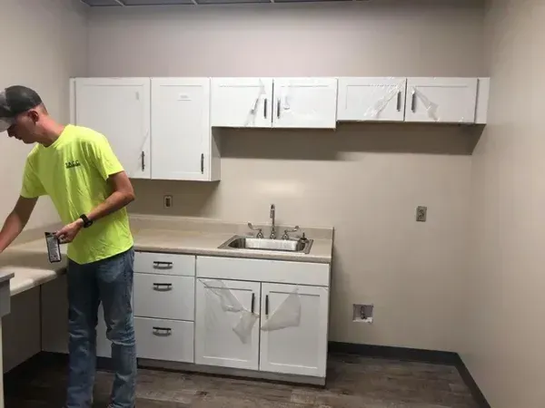 A person in a yellow shirt stands near white cabinets and a sink. Construction setting.