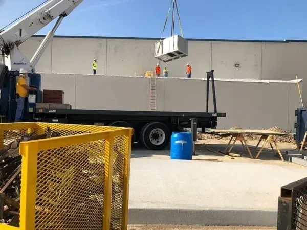 Crane lifting an air conditioning unit; workers in hard hats near a building.