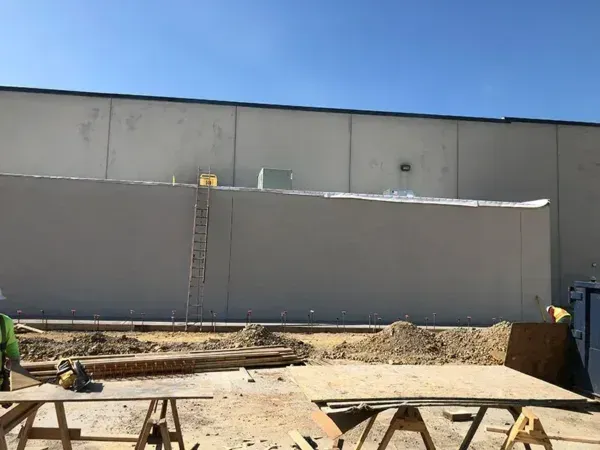 Construction site: workers near a tall gray wall, ladder, and dirt piles under a sunny sky.