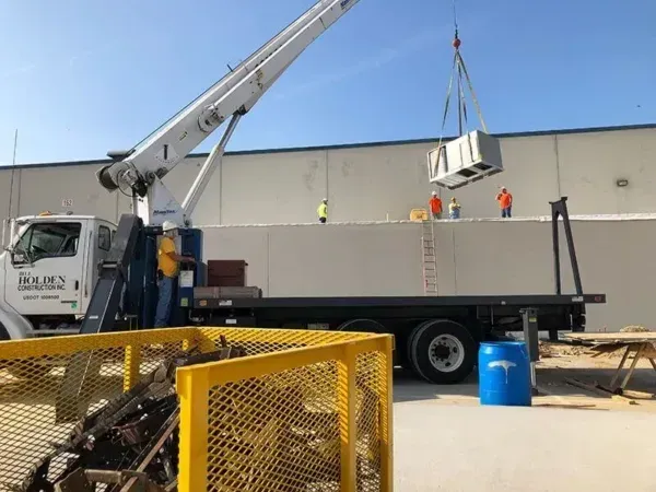 Crane lifting HVAC unit onto a building's roof with workers in orange vests.