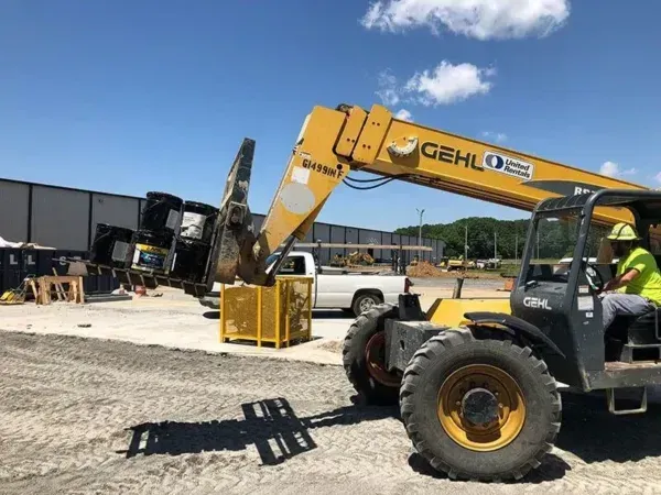 Yellow telehandler lifting a container with construction materials. Operator in cab, blue sky background.