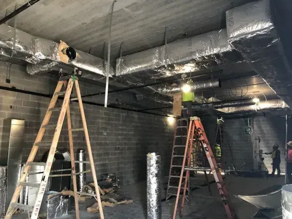 Construction site with workers on ladders installing ductwork. Gray brick walls, concrete ceiling.