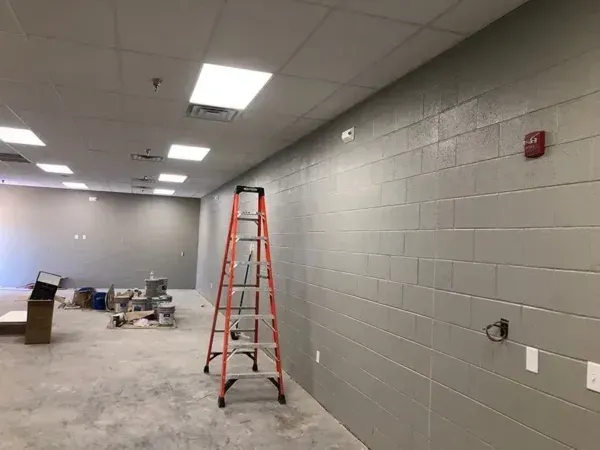 Interior of a room under construction with gray block walls, ceiling lights, a ladder, and paint supplies.
