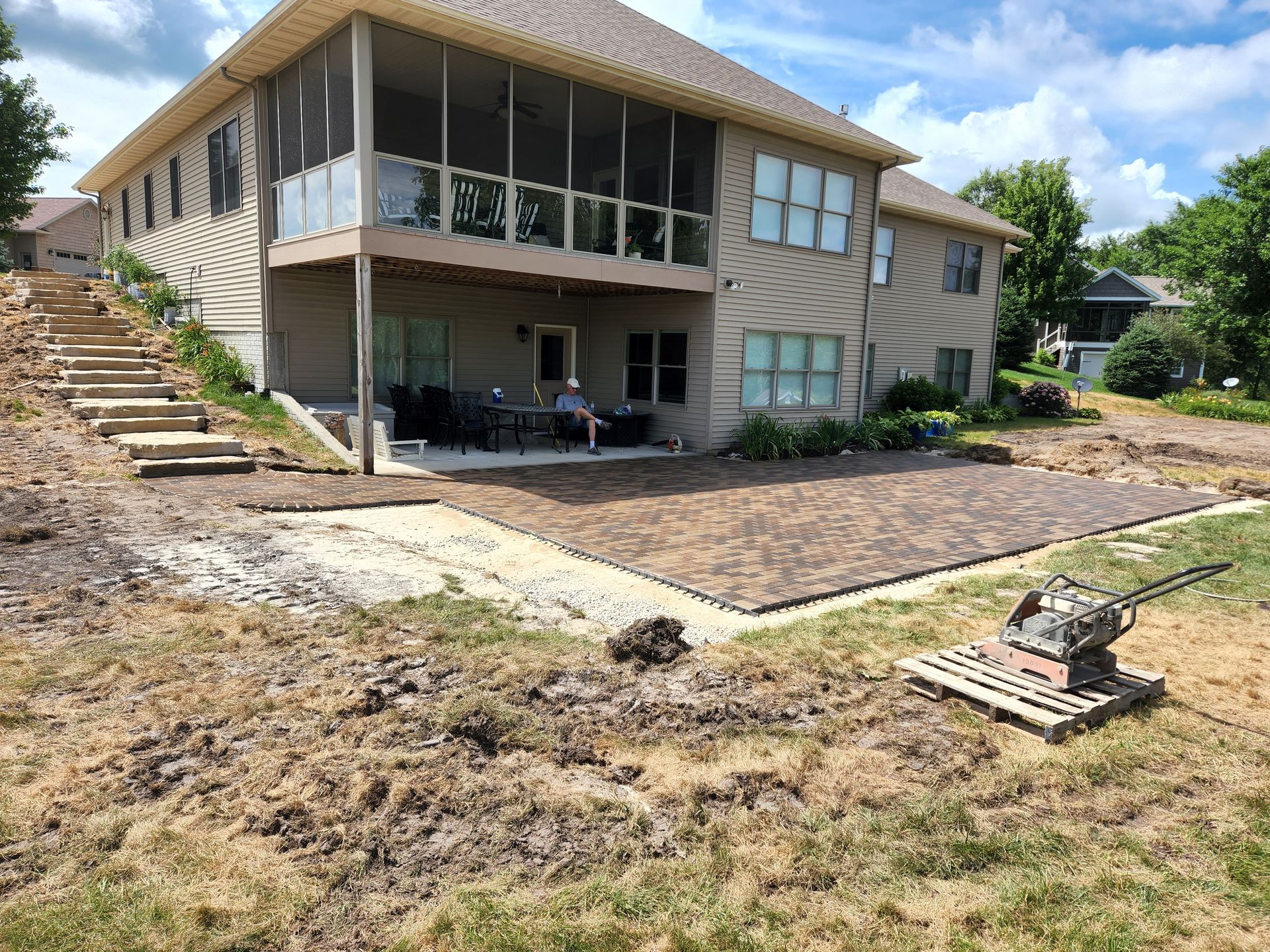 Backyard patio construction in progress; house with screened porch, dirt, pavers, compactor.