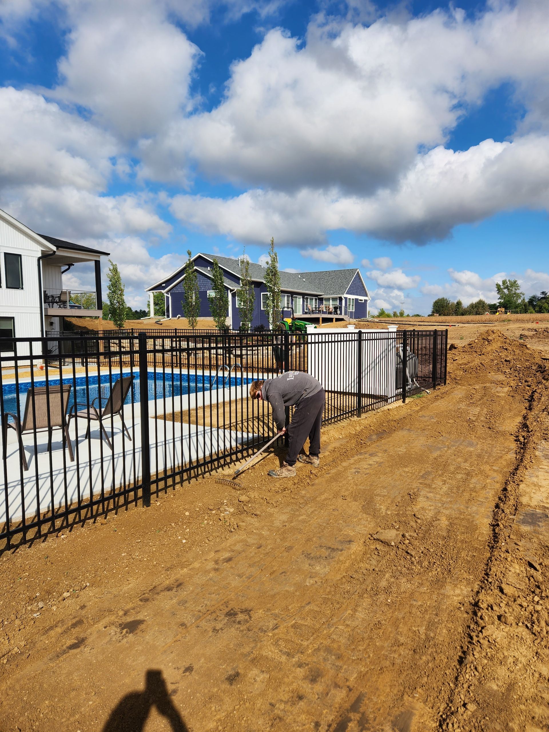 Man working on a dirt path next to a pool and black fence, under a blue sky with fluffy clouds.