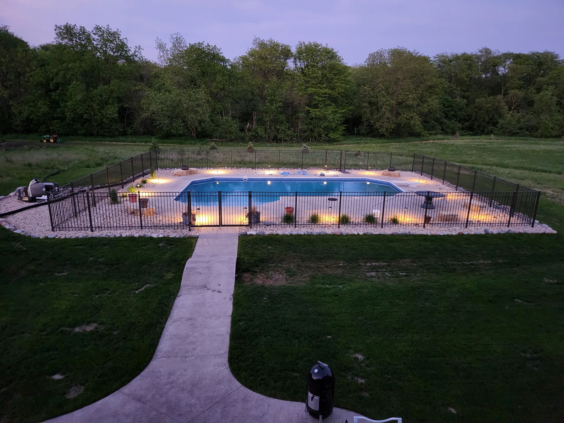 Pool area at dusk, surrounded by black fence and landscaping. Pathway leads to gate.