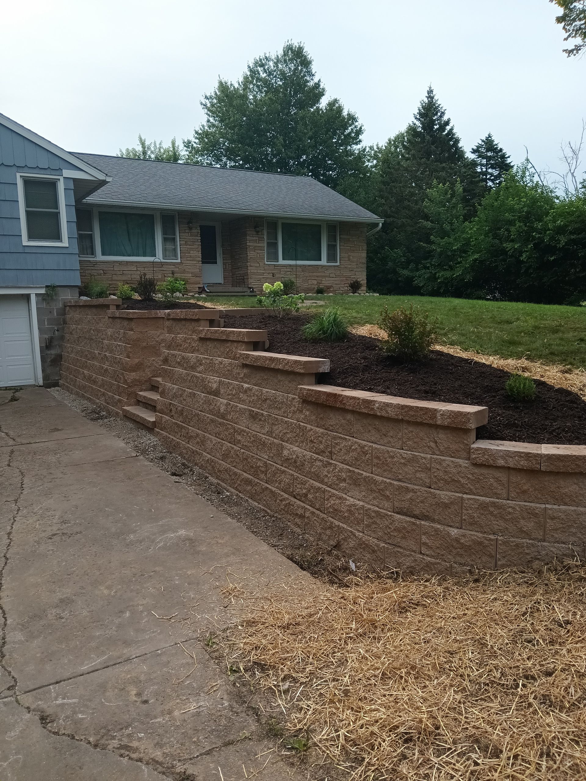House with retaining wall, driveway. Tan brick wall in front, plants on top. Gray roof, blue house.