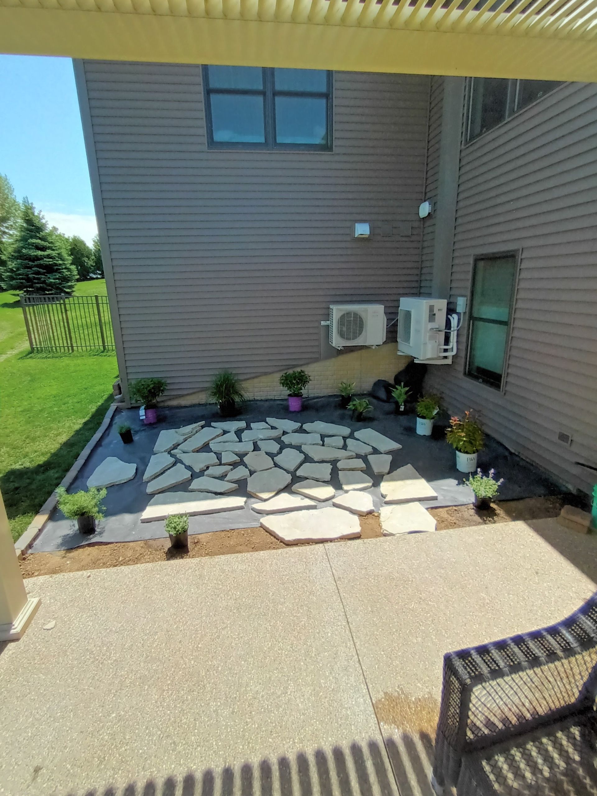 Patio with stone pavers and small plants bordering a house with air conditioning units.