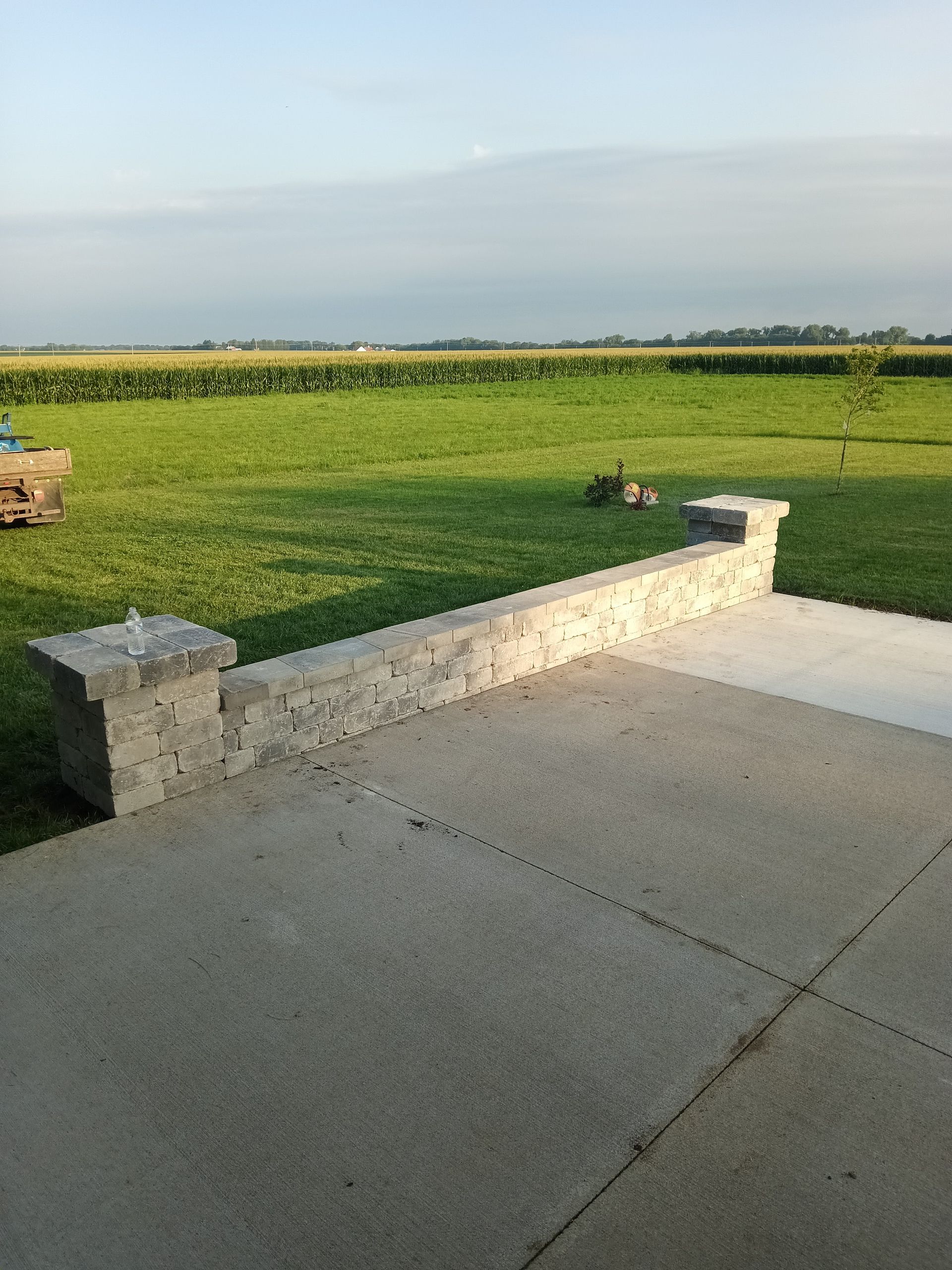 Concrete patio with stone wall overlooking a green field under a cloudy sky.
