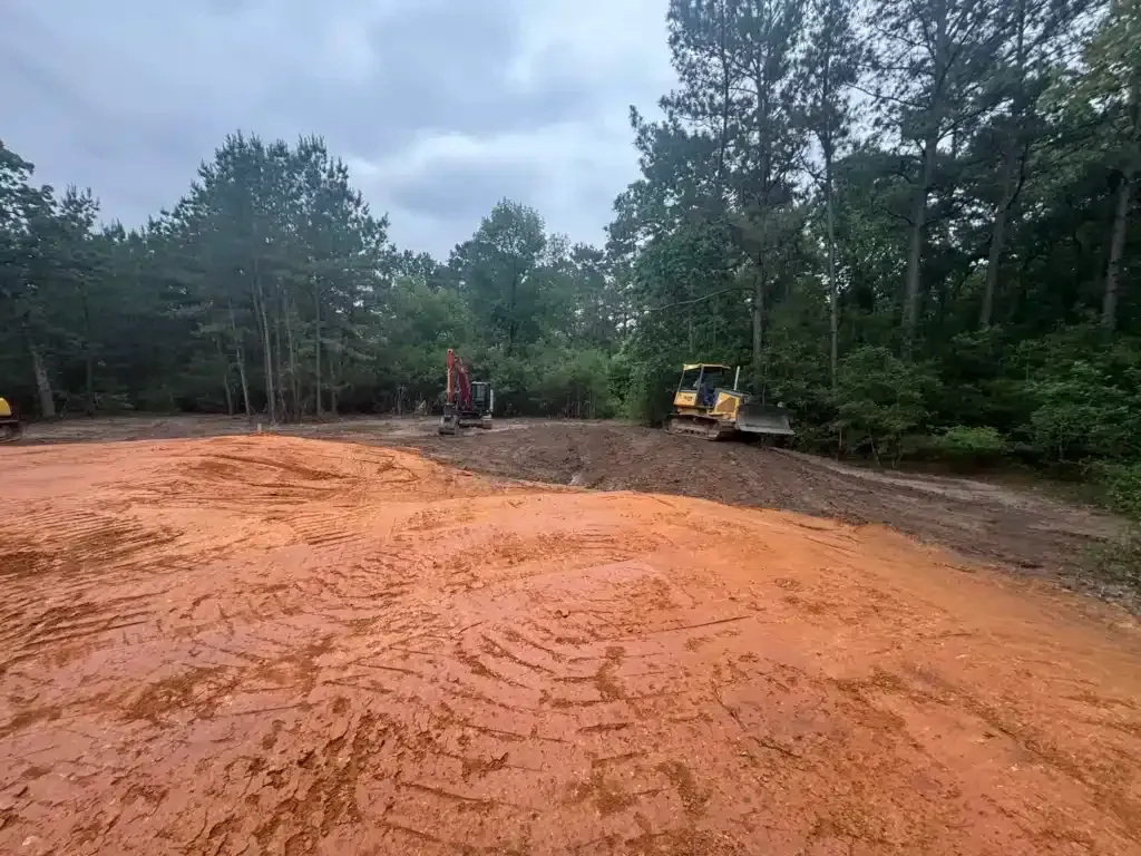 Construction site with orange dirt and two pieces of heavy machinery clearing the land near a forest.