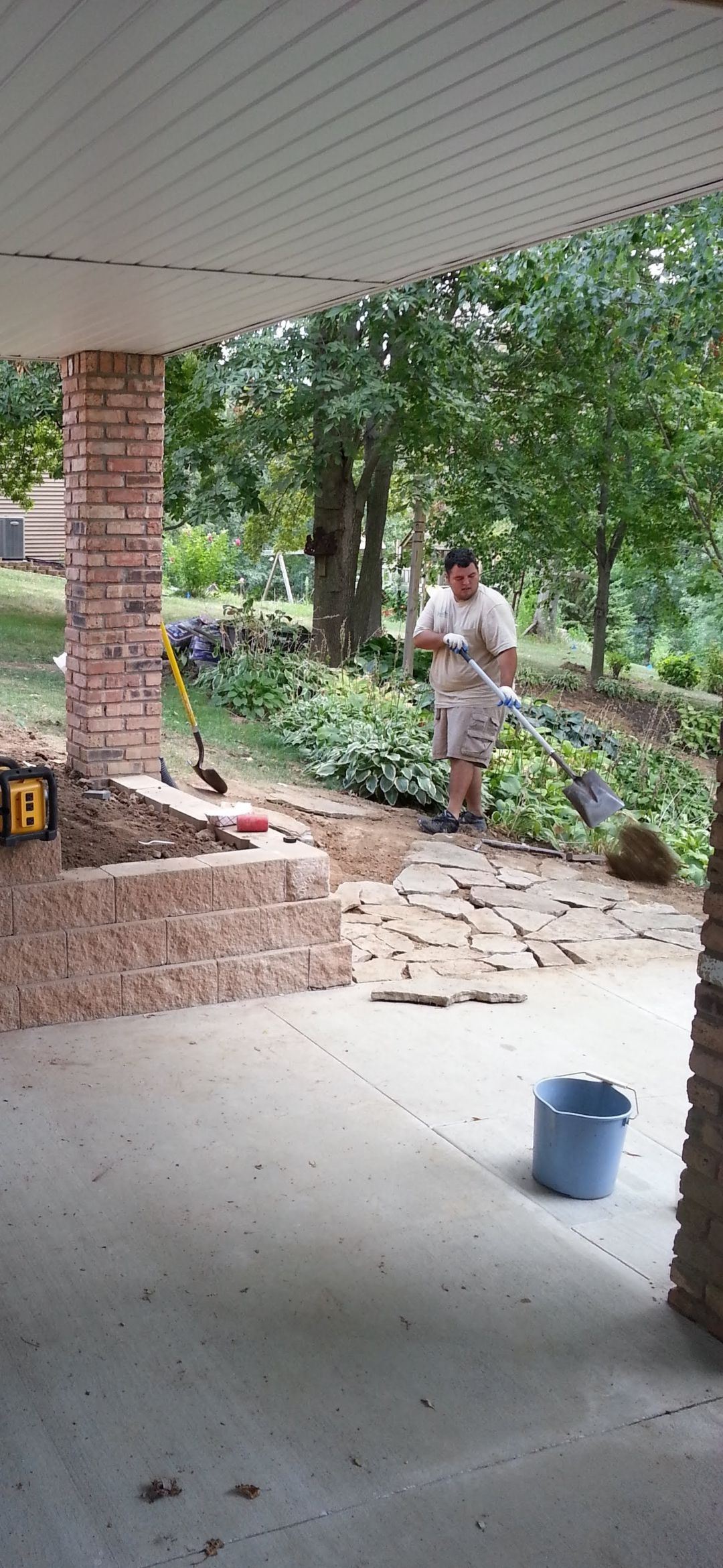 Person using a tool outdoors, near a brick structure. Trees and blue bucket are visible.