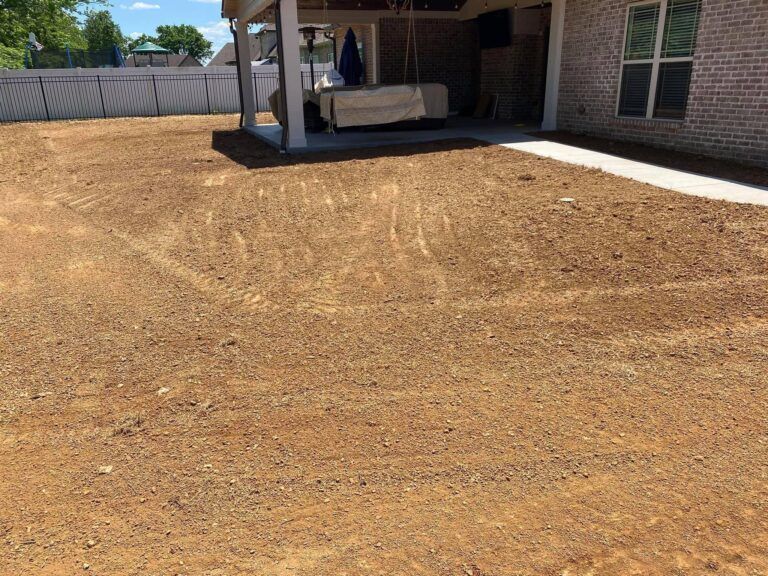 A bare, brown yard with tire tracks, next to a covered patio and brick house under a blue sky.