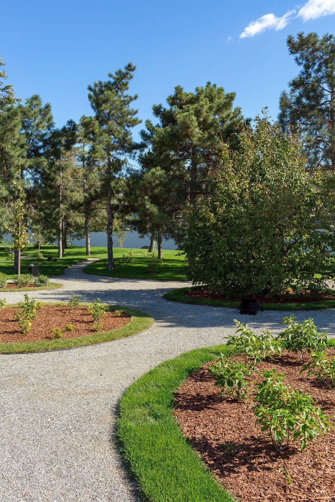 Gravel path winds through a park with trees, grass, and shrubs under a blue sky.