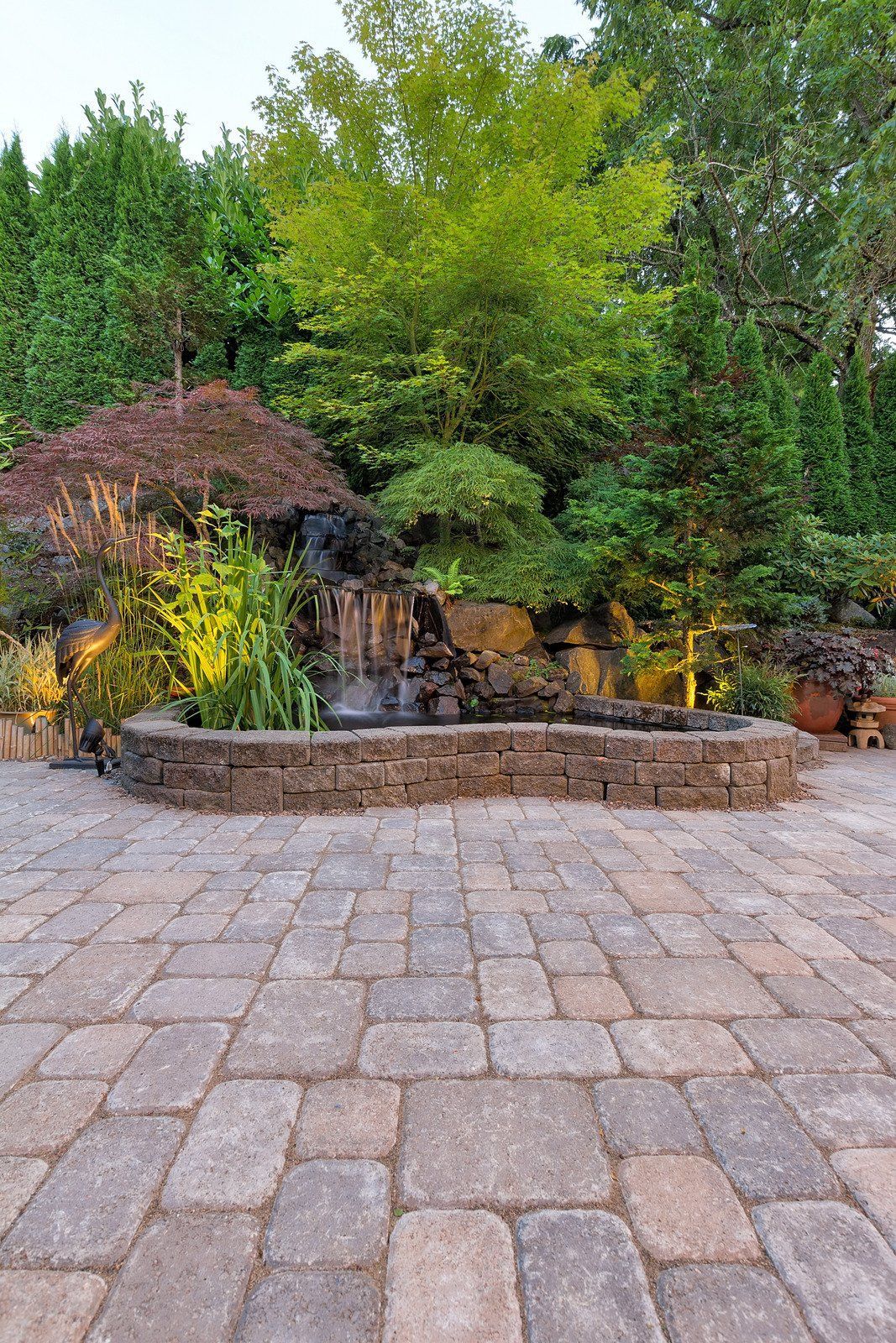 Stone patio leads to a water feature with a waterfall, surrounded by trees and lush greenery.