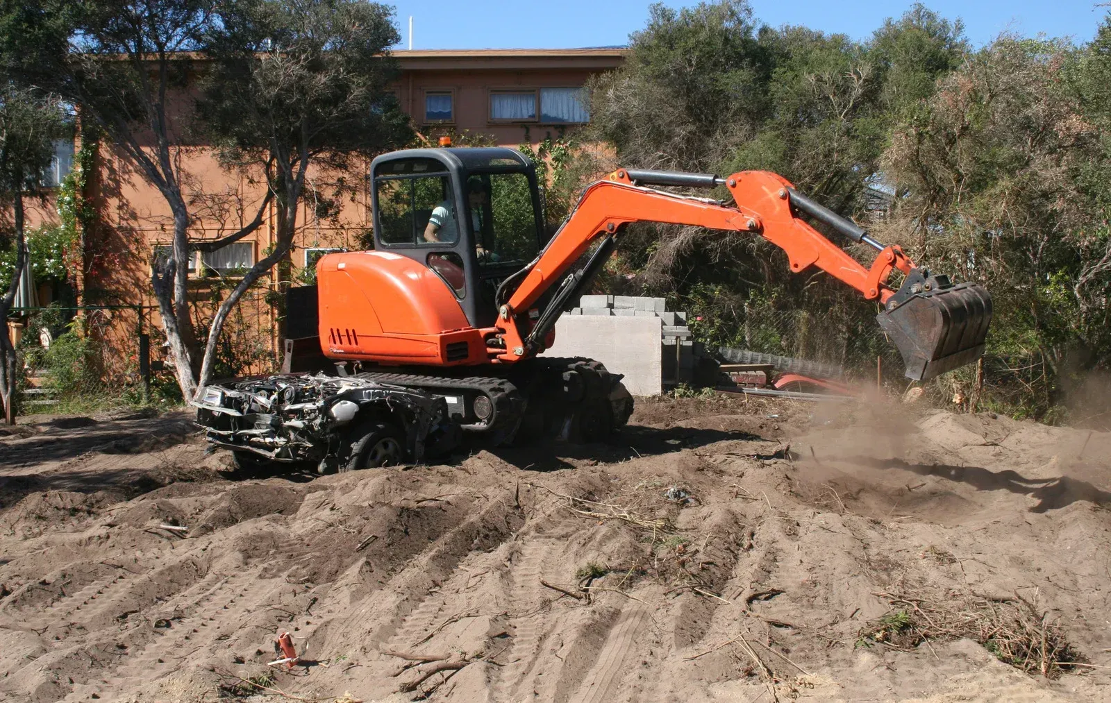 Orange excavator digging in a dirt lot, a building and trees in the background.