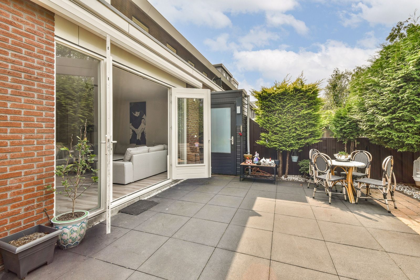 Patio with gray tiles, open doors to living room, brick wall, table, and trees.