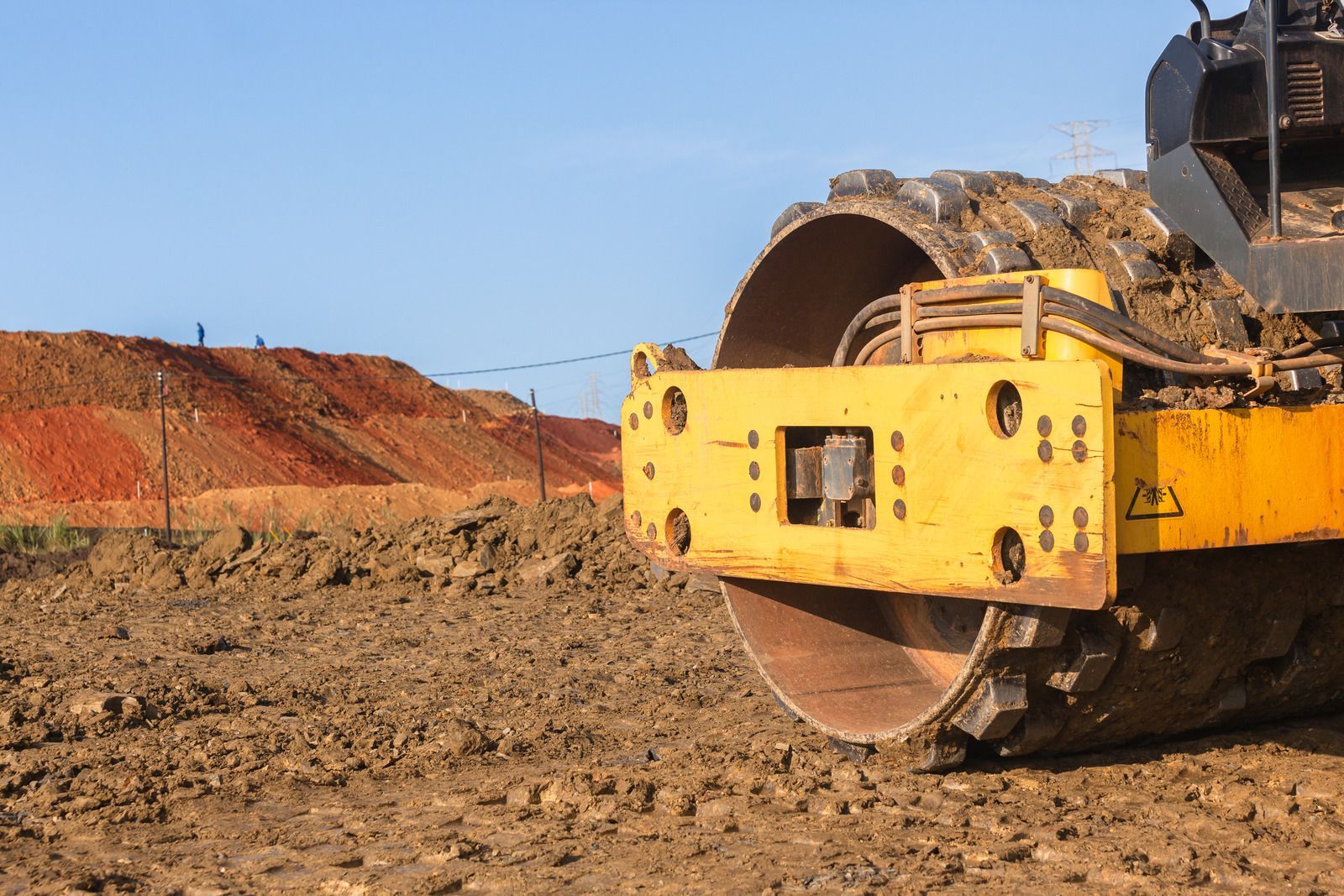 Yellow and black construction roller compacting dirt at a construction site, with a red dirt pile in the background.