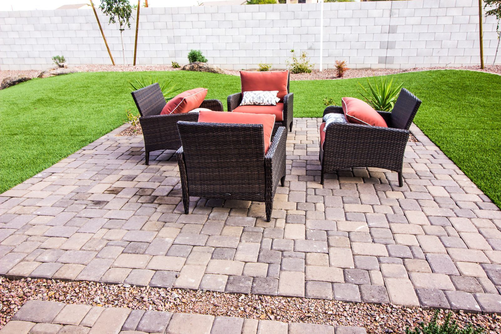 Patio with brick pavers, four wicker chairs with orange cushions, on a lawn with a wall backdrop.